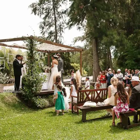 A wedding ceremony held outdoors in a verdant garden, with the bride and groom standing under a wooden arbor, surrounded by guests seated and standing, and children nearby on the grass in Tigre, Buenos Aires.