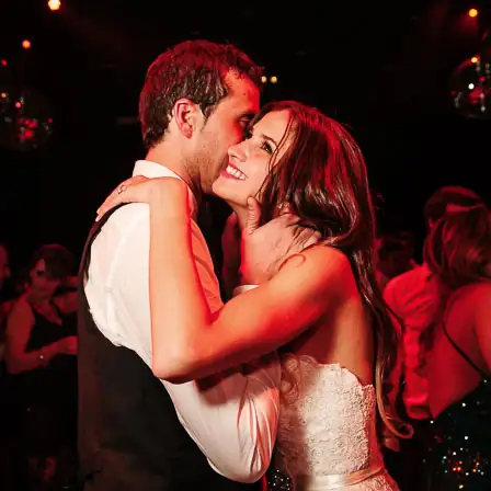 A newlywed couple shares a romantic dance, smiling and embracing each other, with soft lighting and blurred guests in the background at a wedding reception in Buenos Aires.
